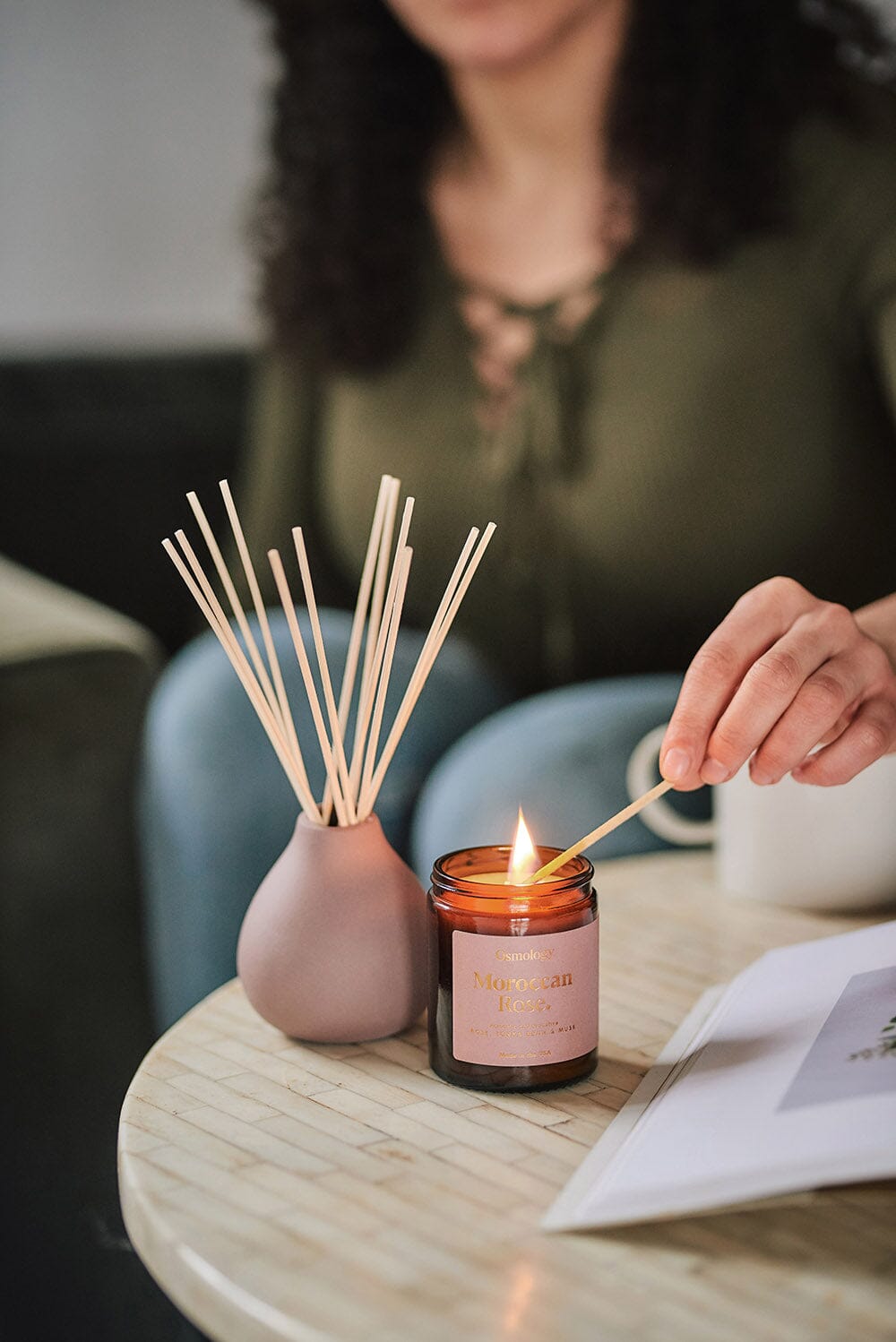 Osmology Moroccan Rose jar candle being lit by person holding match with open lid with Moroccan Rose reed diffuser on coffee table