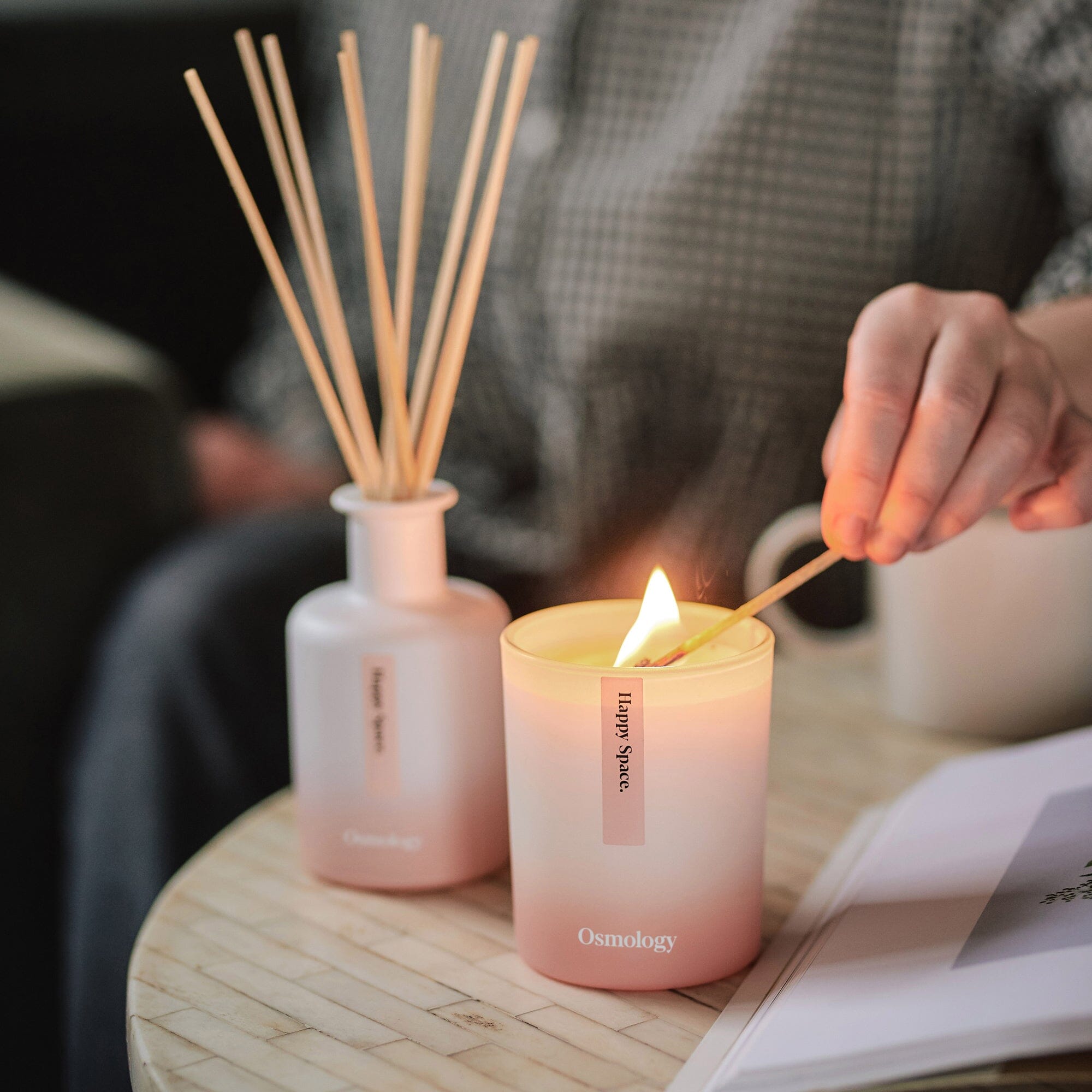 pink happy space candle and diffuser by osmology displayed on coffee table with person lighting candle