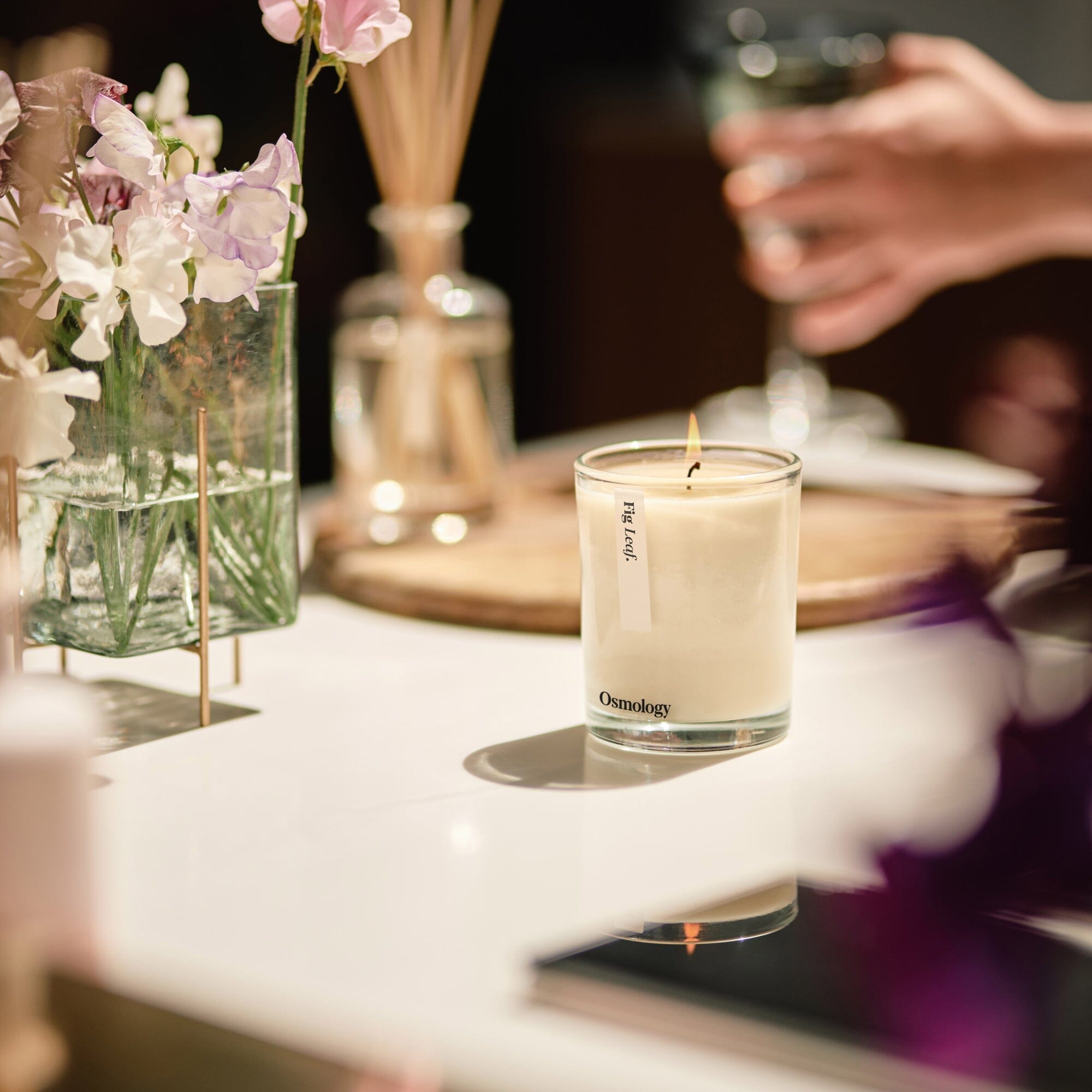 lit osmology fig scented candle displayed in a sunny kitchen setting next to sweet pea flowers