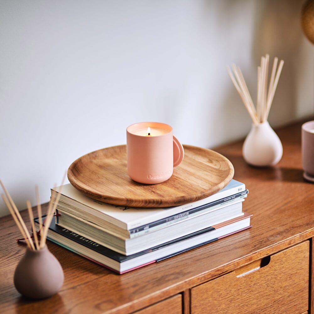 Osmology Aztec Tuberose candle, Peach coloured ceramic candle with lit flame and matching ceramic lid on wooden tray