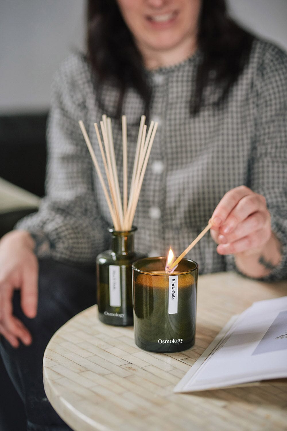 Osmology Black Oak scented candle being lit by match on coffee table, Black Oak diffuser behind the candle out of focus