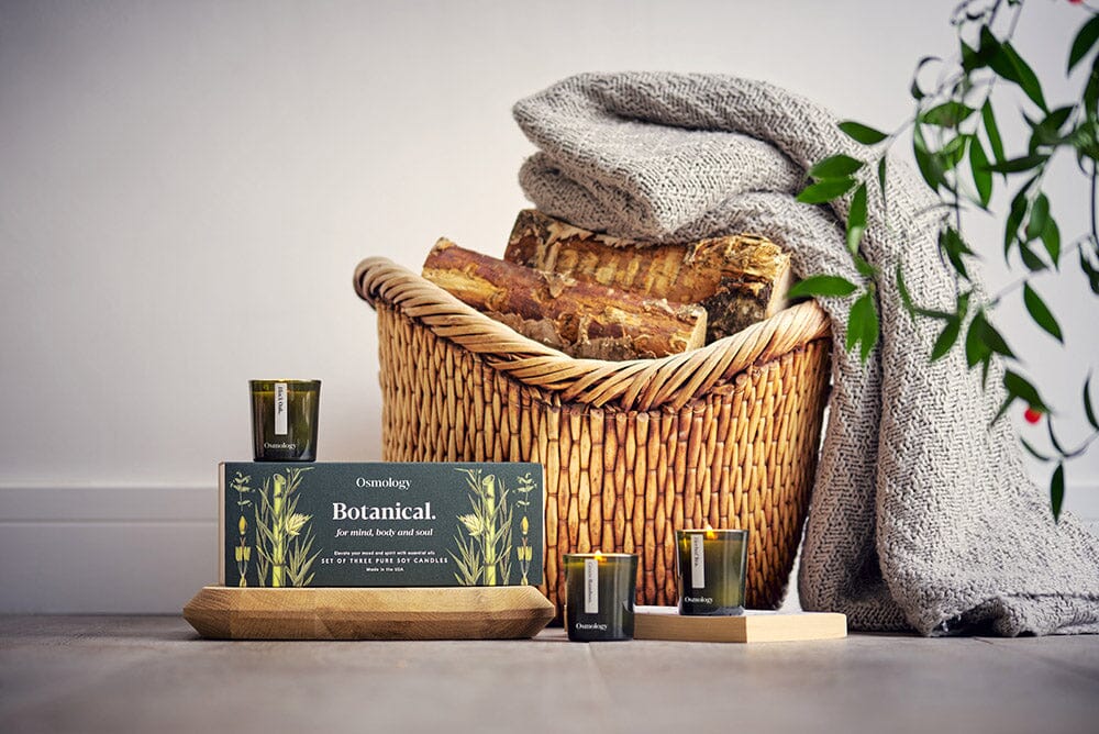 Osmology Botanical gift set, with three candles, arranged on a wooden board and on floor, a basket of logs with a blanket draped over it in background, out of focus plant leaf textures