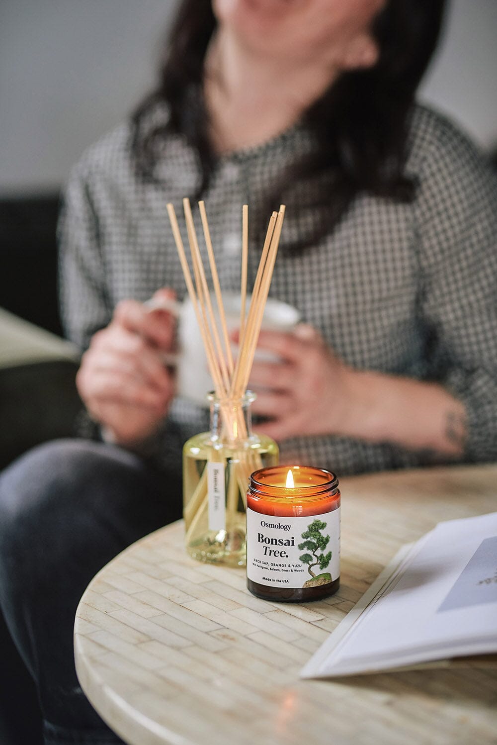 Osmology Bonsai Tree jar candle with lit candle flame on coffee table with Bonsai Tree diffuser behind, person holding mug in background