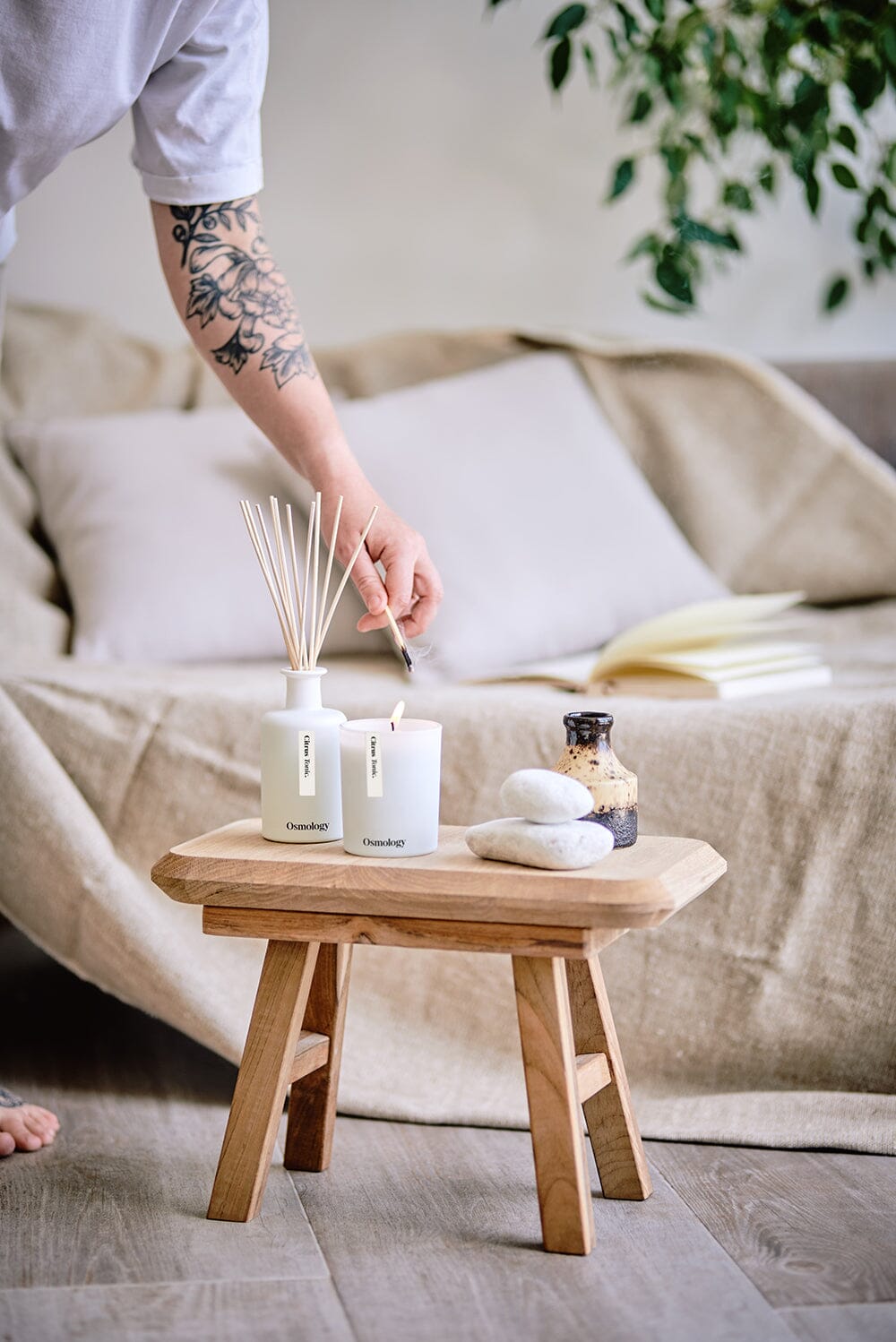 A person lighting Osmology Citrus Tonic candle with match, Citrus Tonic diffuser and decorative stones placed next to it on wooden stand