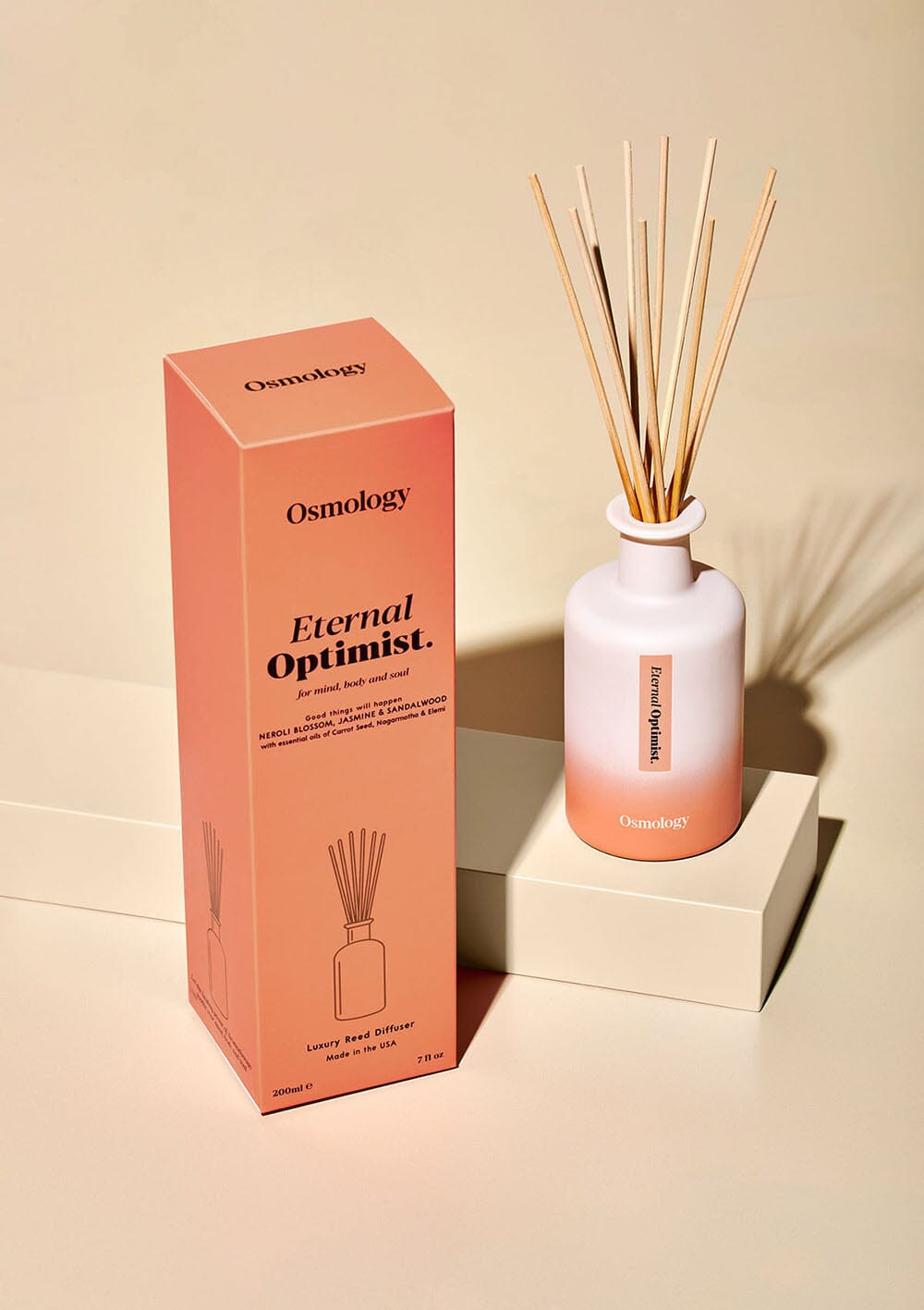 Osmology Eternal Optimist reed diffuser on plinth next to product packaging, cream background