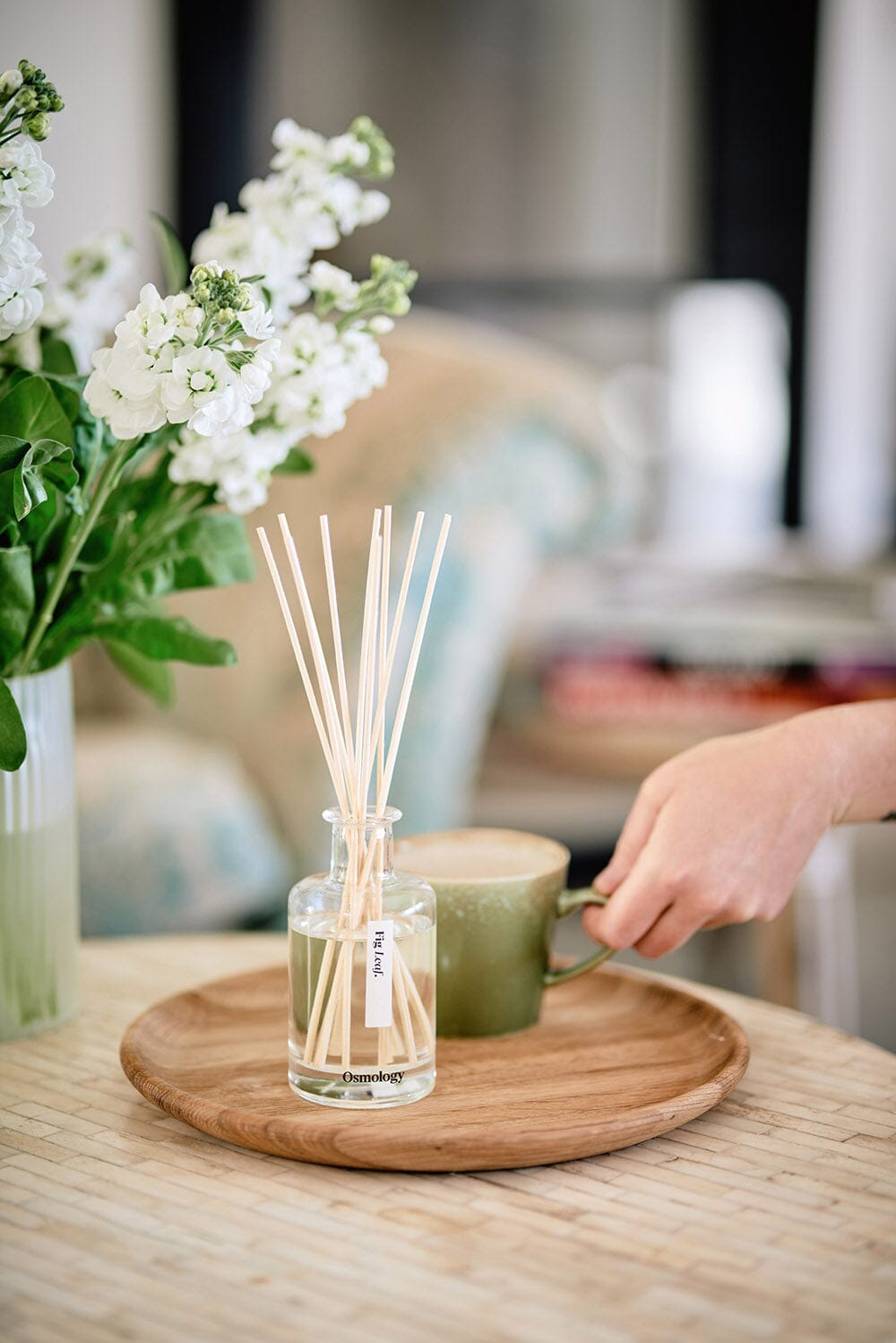 Osmology Fig Leaf diffuser on wooden tray with a hand holding a mug, flowers  