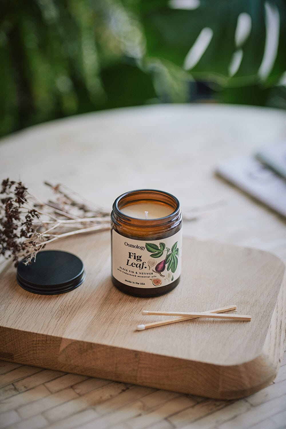 Osmology Fig Leaf jar candle with lid placed next to it, two matches balanced on top of each other in front, all arranged on wooden board