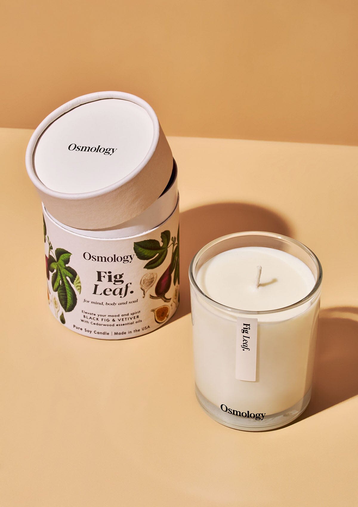 Osmology Fig Leaf candle with decorative product packaging with lid slightly ajar, orange-cream background