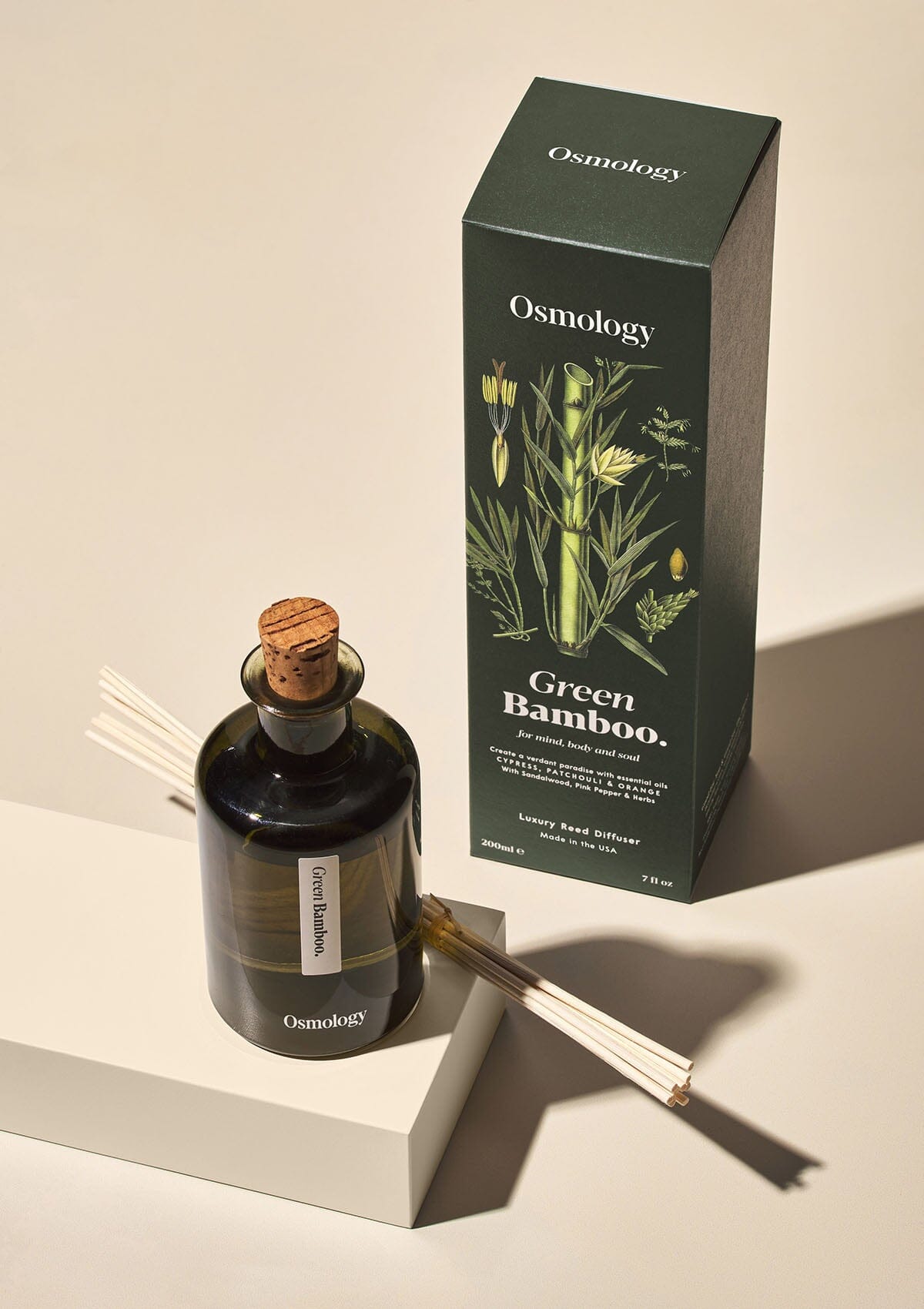 Osmology Green Bamboo reed diffuser with cork stopper in and bunched up reeds balanced on a plinth, product packaging behind, cream background