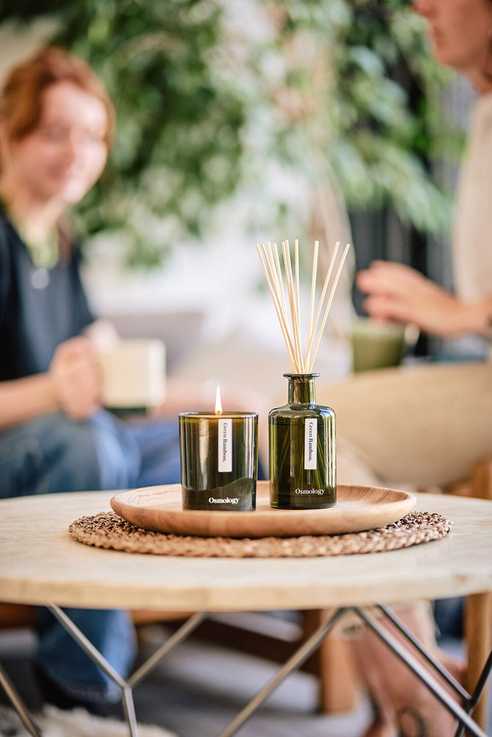 Osmology Green Bamboo reed diffuser next to  Green Bamboo candle with flame on wooden tray, arranged on coffee table, two women sat drinking coffee/tea in background  