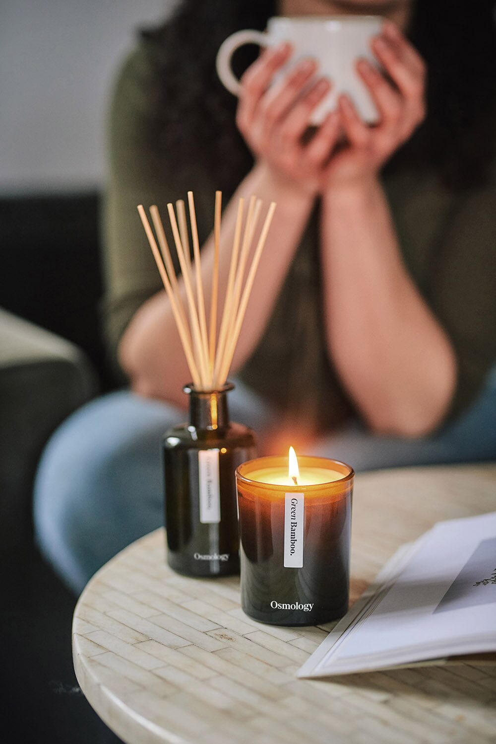 Osmology Green Bamboo candle with lit flame, Green Bamboo reed diffuser behind, arranged on coffee table with open book, person in background holding mug up with two hands