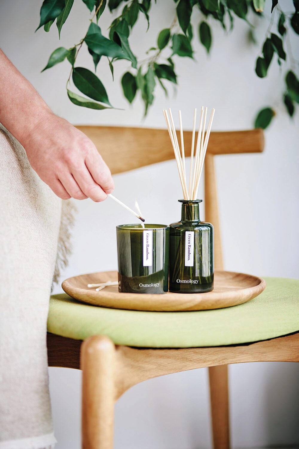 Osmology Green Bamboo candle being lit by person holding a match, Green Bamboo reed diffuser placed next to it on wooden tray that is arranged onto a chair, blanket draped over chair