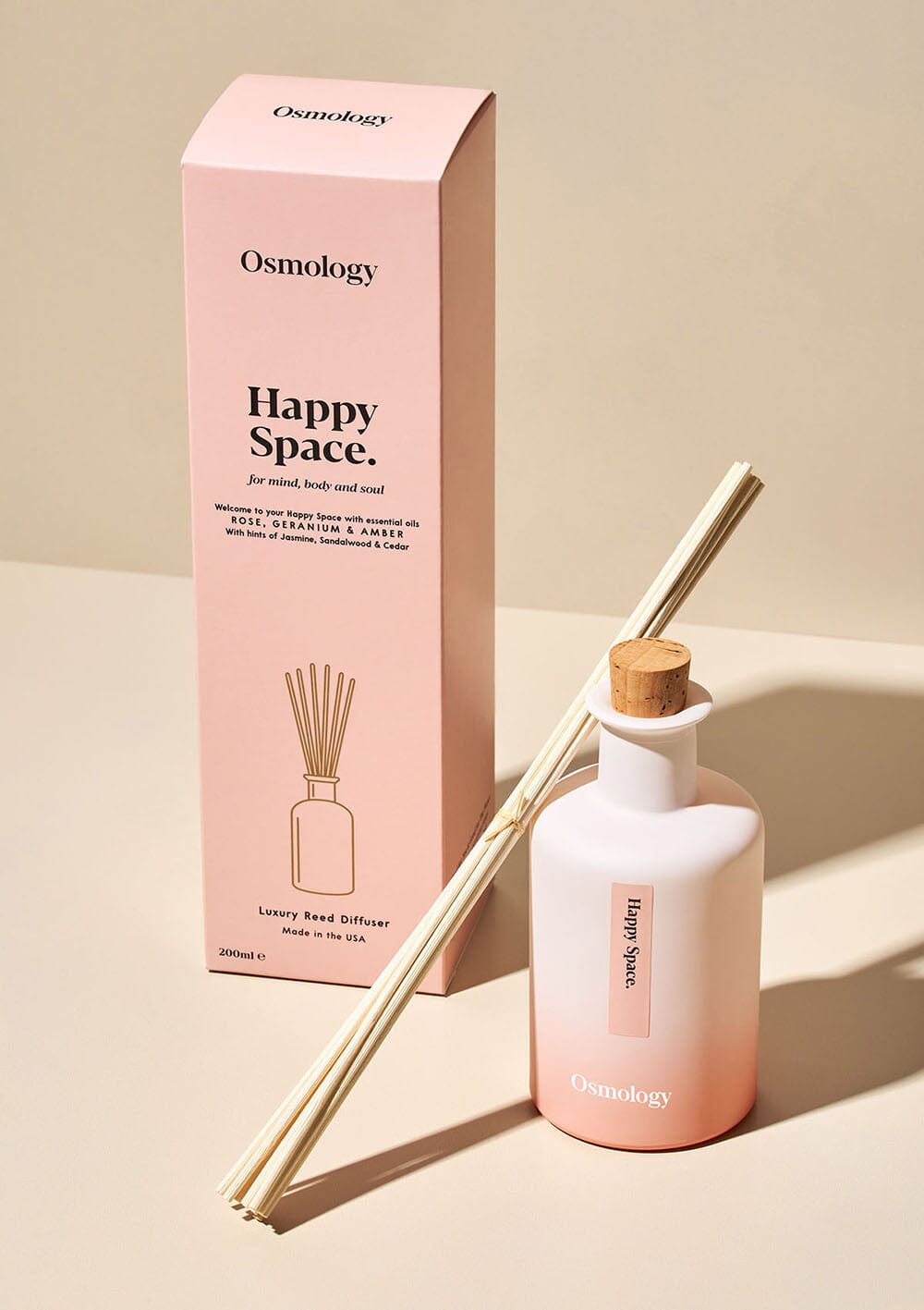 Osmology Happy Space reed diffuser with cork stopper in and a bunch of reeds leaning against it, product packaging behind, on cream background