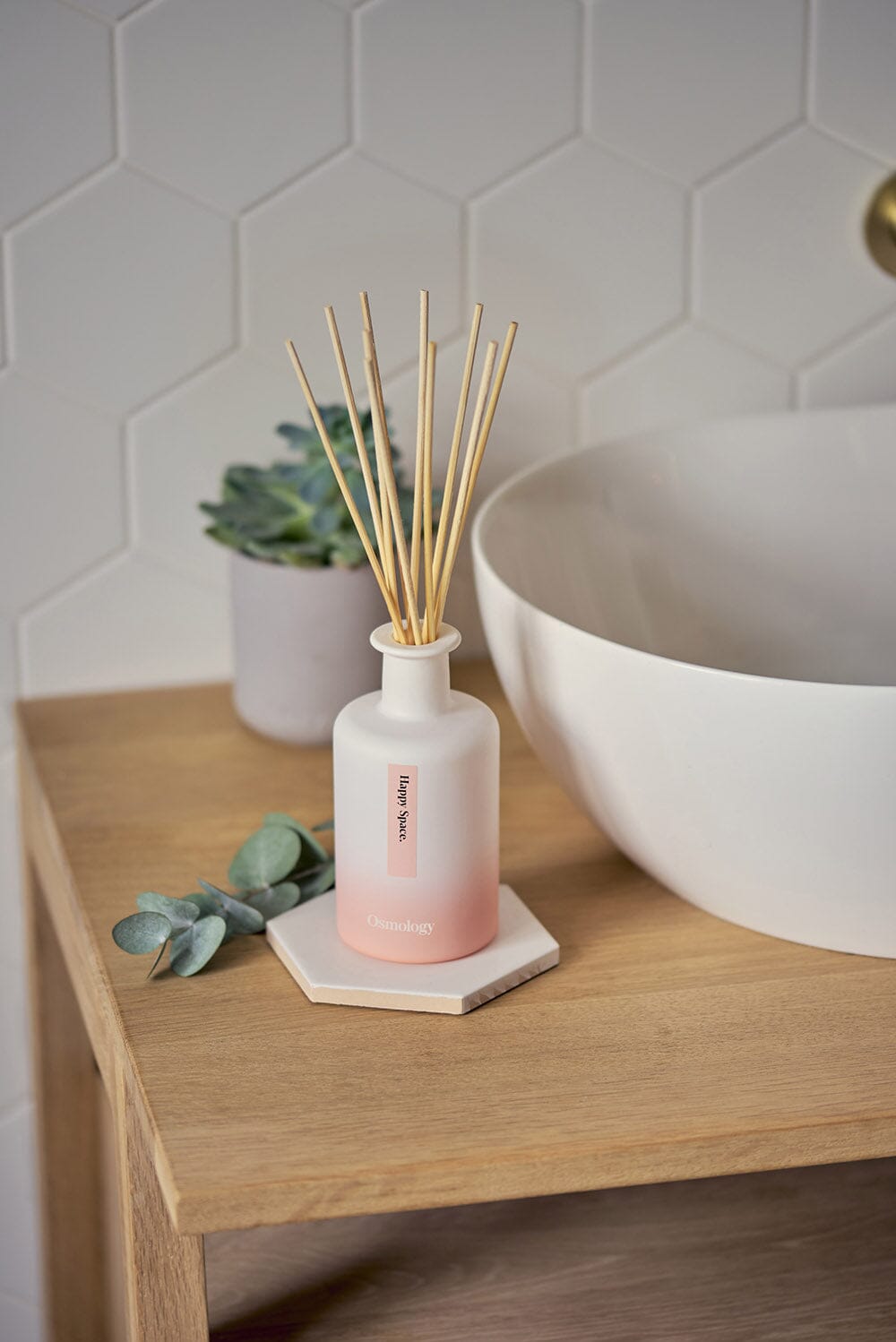 Osmology Happy Space reed diffuser placed on hexagon tile, next to stylish sink with wooden cabinet, stylish bathroom setting