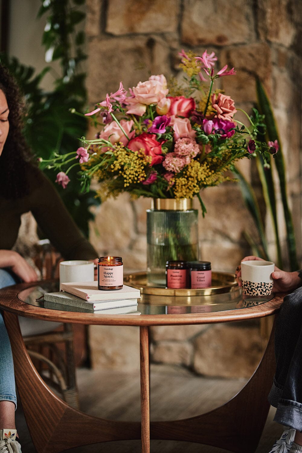 A coffee table with two people sat at, Osmology Happy Space jar candle with lit candle flame placed on books,  colourful flowers in vase on table, two other jar candles on table behind