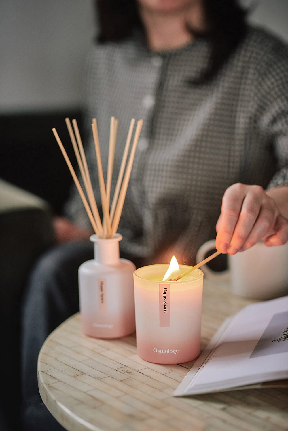 Osmology Happy Space candle being lit with match by person, Happy Space diffuser out of focus next to candle