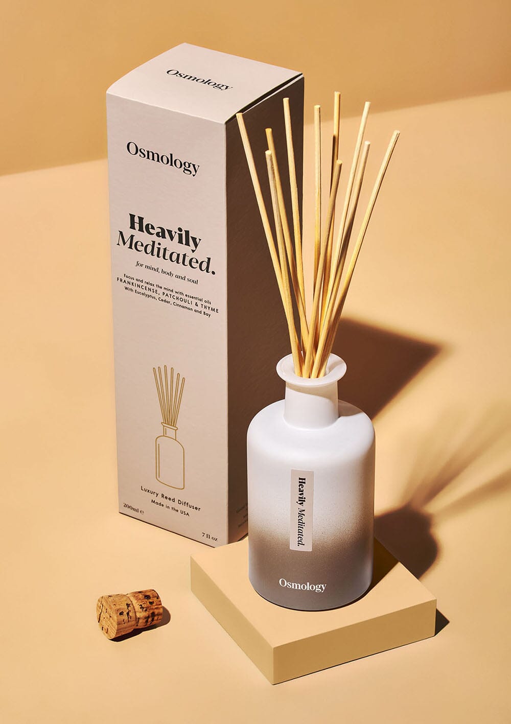 Osmology heavilly meditated reed diffuser next to product packaging, cork on floor, all on an orange background