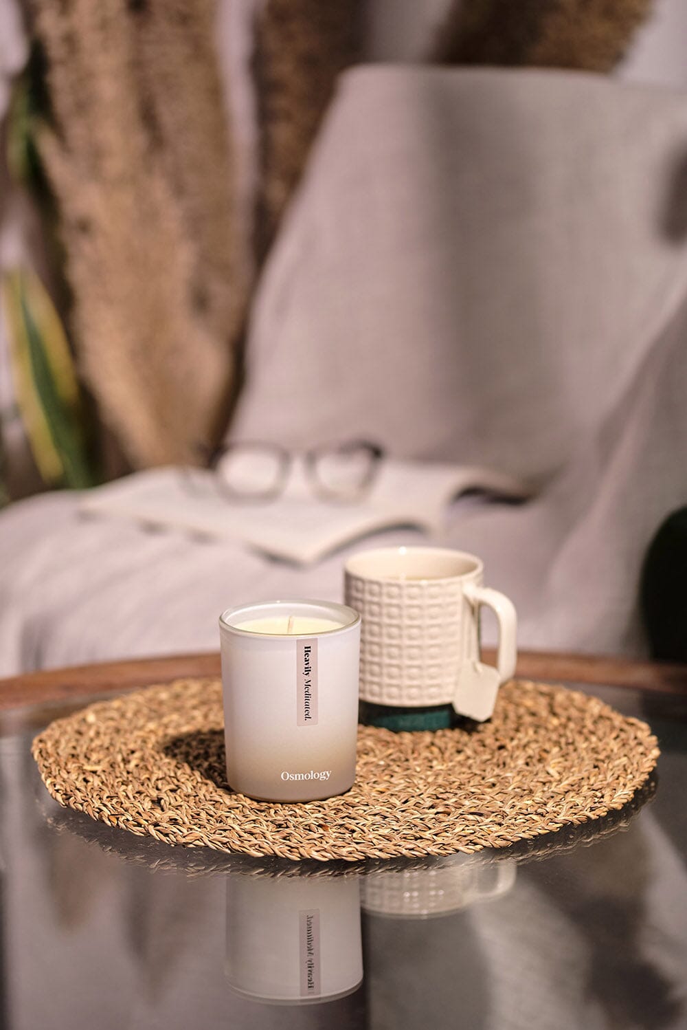 Osmology Heavily Meditated candle on glass coffee table with mug behind, reflected in glass below, book with glasses behind 