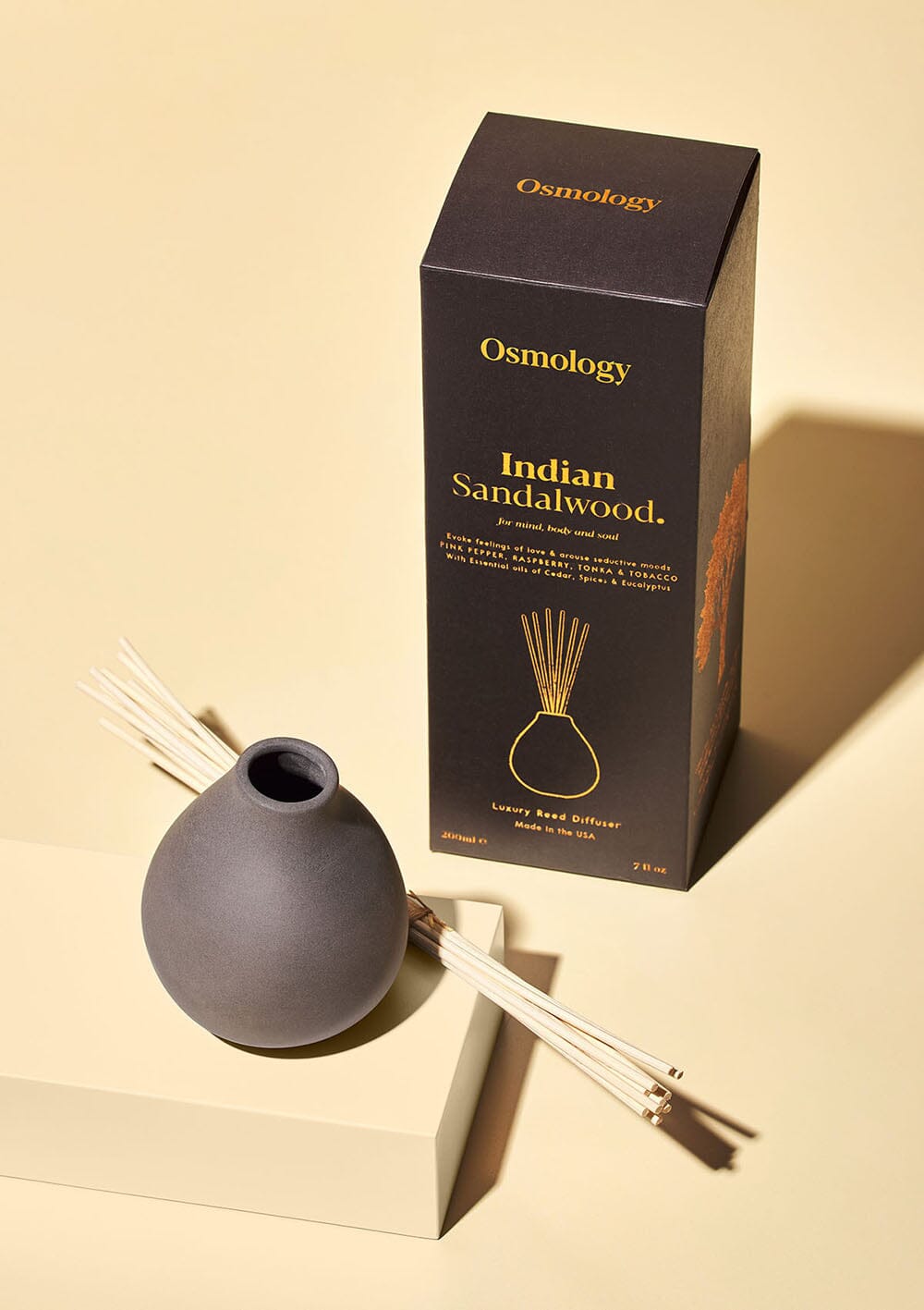 Osmology diffuser pot and bunched up reeds next to product packaging on a yellow-cream background