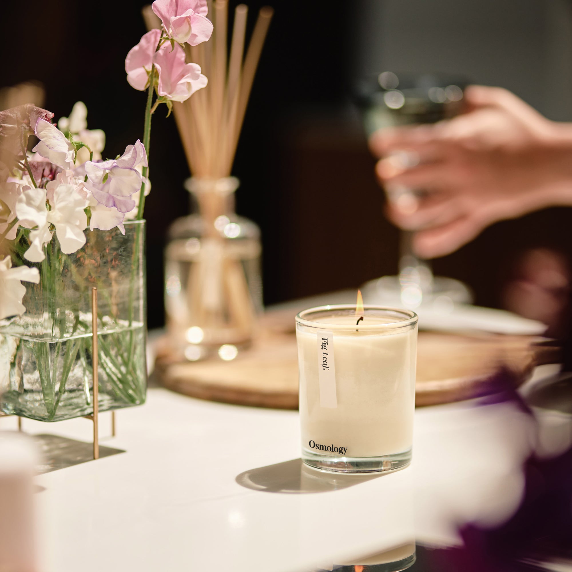 lit osmology fig scented candle displayed in a sunny kitchen setting next to sweet pea flowers