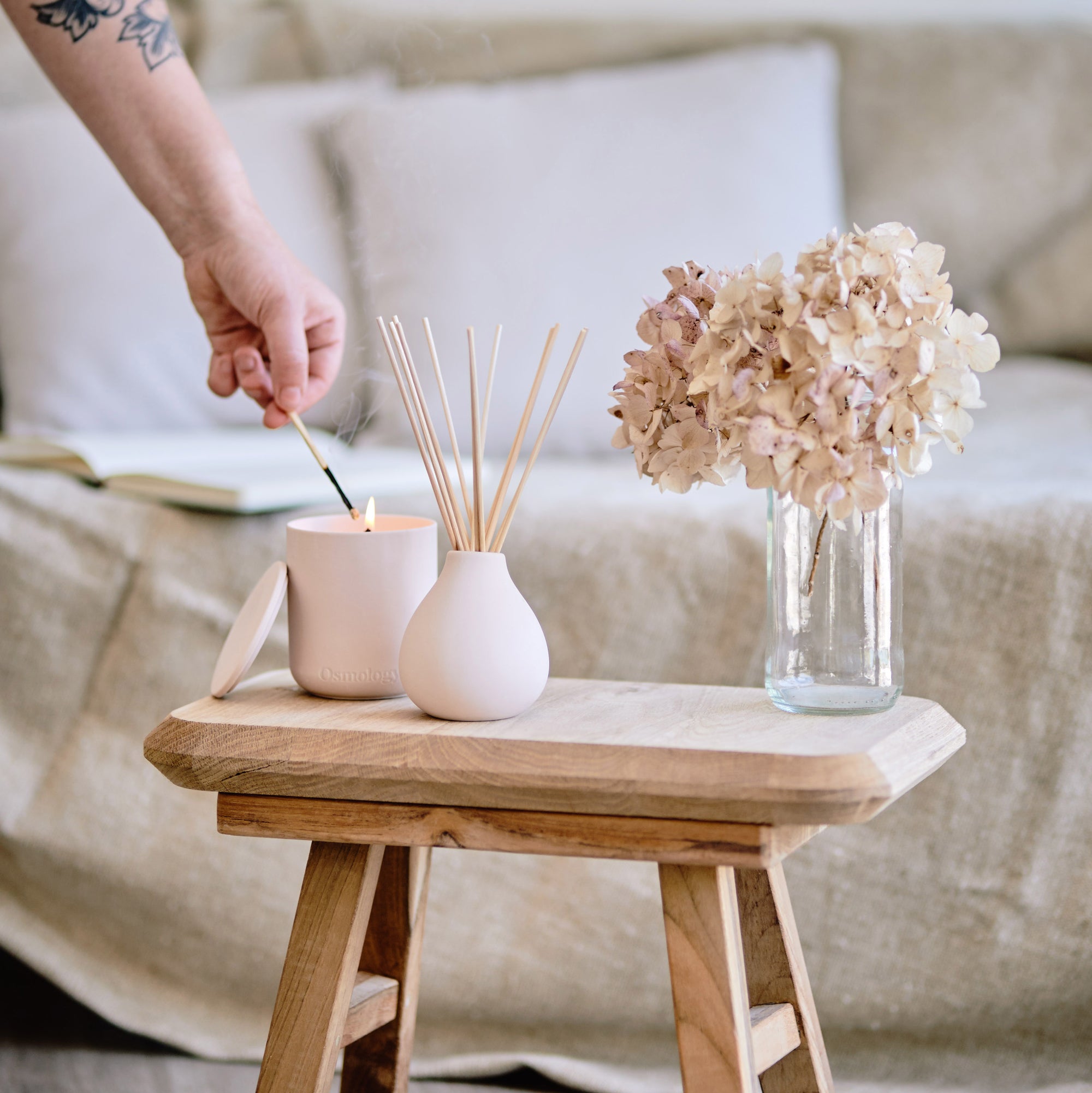 Person lighting pink ceramic candle displayed next to matching diffuser on small coffee table next to some dried decorative flowers