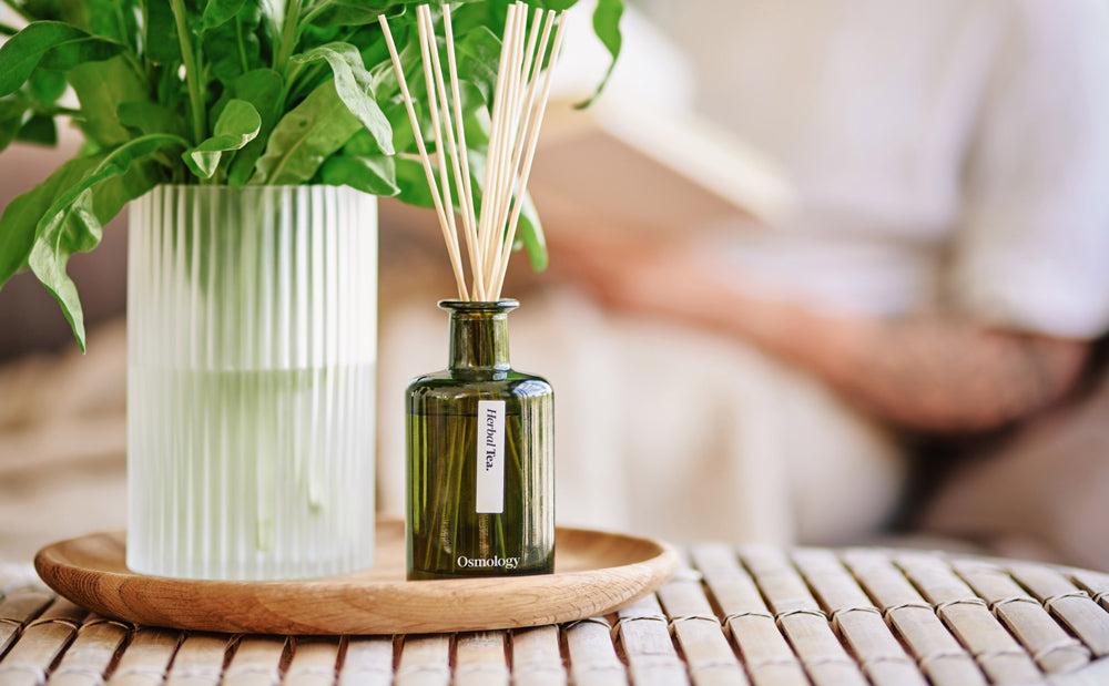 green glass reed diffuser displayed on wooden tray on a coffee table with person reading in the background