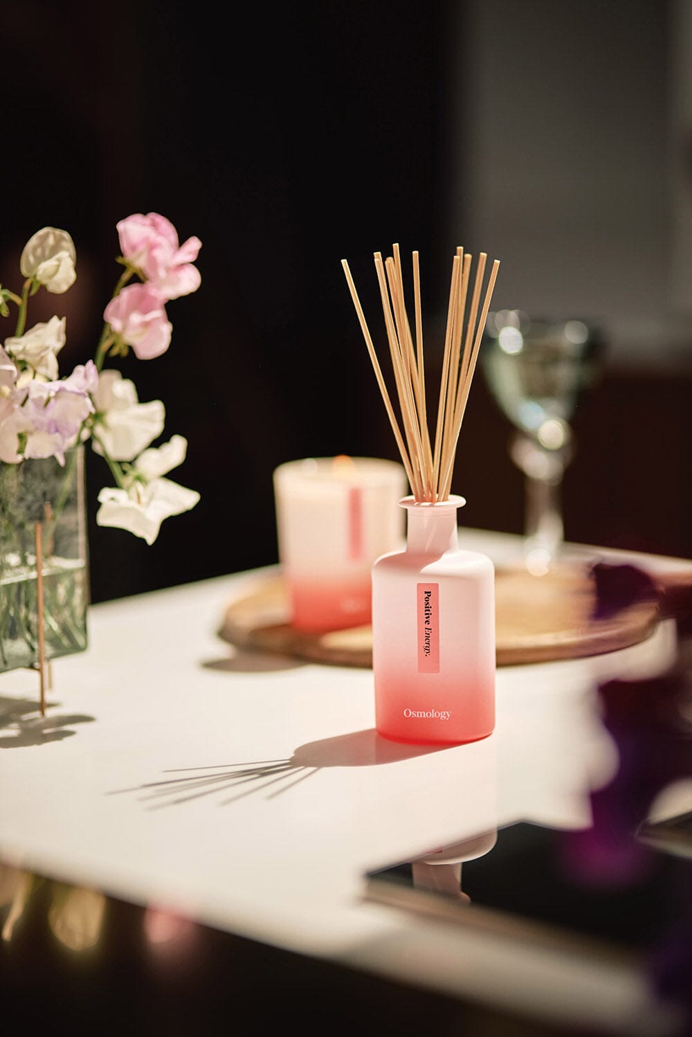 Osmology Positive Energy Diffuser on stylish table, with wooden tray and candle and in background, flowers and glass  